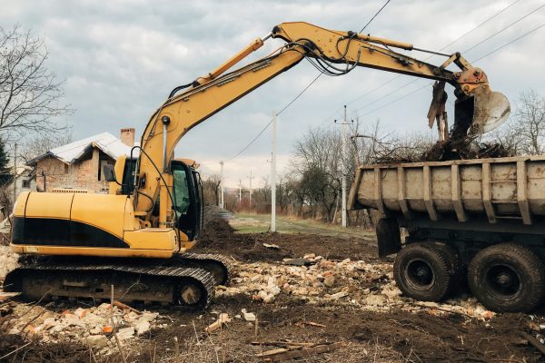 Bulldozer clearing land from old bricks and concrete from walls with dirt and trash. Backhoe machinery ruining house. Excavator destroying brick house on land in countryside.